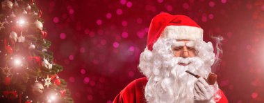 Noel Baba Noel ağacının önünde pipo içiyor, kırmızı arka planda.
