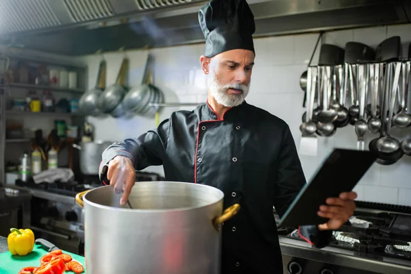 Mature chef stirs a pot while consulting a digital tablet in a commercial kitchen