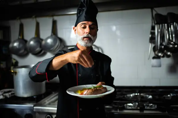 Mature chef in uniform focuses on garnishing a plated meal in a commercial kitchen setting