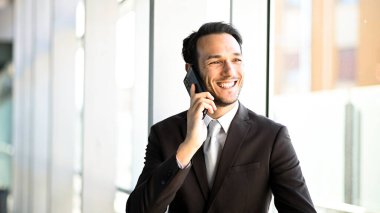 Takım elbiseli neşeli erkek profesyonel modern ofis ortamında cep telefonuyla sohbet ediyor.