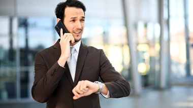 Takım elbiseli profesyonel bir adam, dışarıdaki bir telefon görüşmesinde saatini kontrol ederek birden fazla iş yapıyor.