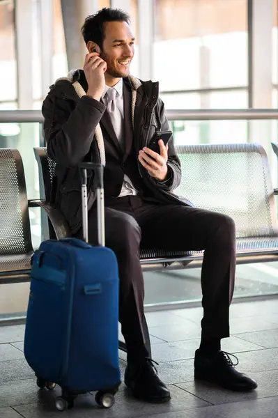 Cheerful young man chats on his mobile phone while waiting with luggage at the airport terminal