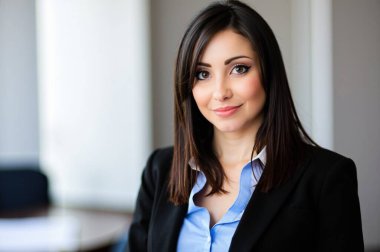 Genç iş kadını, modern bir ofis binasında siyah ceket ve mavi gömlek giyerek poz veriyor. Gülümsüyor ve kendinden emin görünüyor.