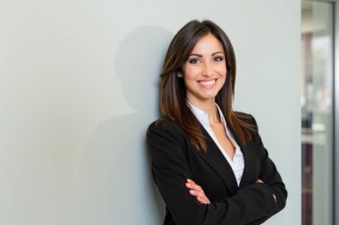 Şık ofis ortamında kendine güvenen bir iş kadını. Kollarını kavuşturup gülümseyerek liderlik ediyor. Başarı ve mutluluk saçıyor.