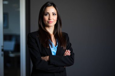 İş kadını kollarını kavuşturarak bir ofiste duruyor, profesyonellik, güven ve kararlılık hissi veriyor.