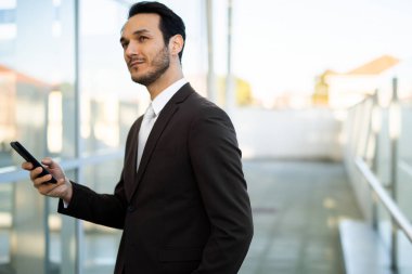 Elinde telefon tutan takım elbiseli genç bir adam, düşünceli bir şekilde şehir ortamında yüzünü çeviriyor.