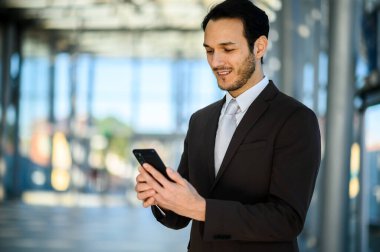 Modern şehir ofis ortamında e-posta ve mesajları kontrol etmek için akıllı telefon kullanırken gülümseyen genç profesyonel işadamı