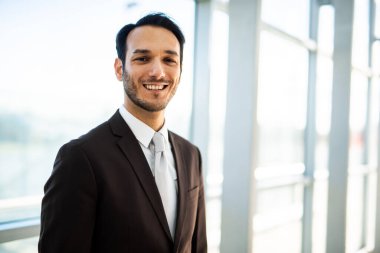Modern bir ofis binasında gülümseyen genç bir işadamı, bir şirket portresi için güven ve başarı yayıyor. Parlak iç yapısı, iş dünyasının lideri olarak mesleki tavrını yansıtıyor.