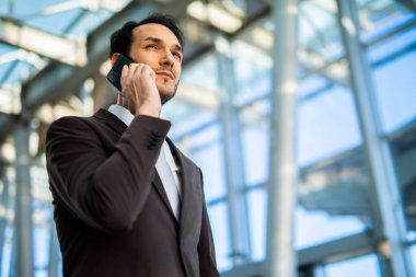 İş adamı, geniş pencereli modern bir ofis binasında telefon görüşmesi yapıyor. O bir takım elbise ve kravat takıyor ve kameradan uzağa bakıyor.