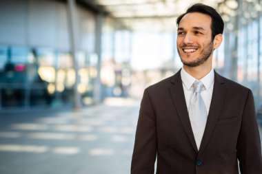 Genç işadamı ofis binasının dışında yürürken gülümsüyor. Kameraya bakmıyor ve mutlu görünüyor.