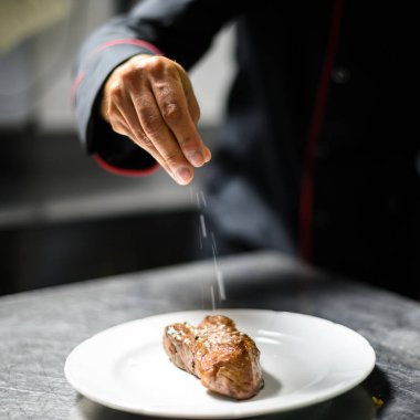 Şef lezzetli bir biftek tabağına son dokunuşları ekler, beyaz bir tabağa tuz serper. Uzman elleri iş başında yakalar, profesyonel bir mutfakta mutfak sanatını gözler önüne serer