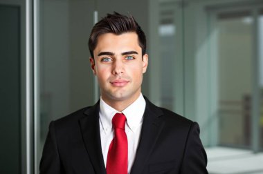 Kendine güvenen, şık takım elbiseli, kırmızı kravatlı, modern ofis ortamında gülümseyen genç bir iş adamı. Kurumsal lider olarak başarı ve profesyonellik yayar.
