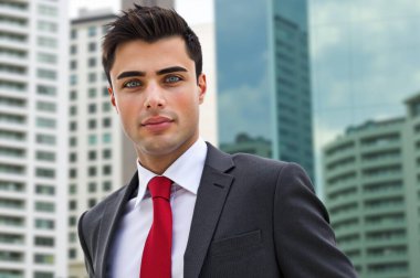 Kırmızı kravatlı kendine güvenen genç işadamı şehir manzarasında poz veriyor, başarı ve liderlik yayıyor.