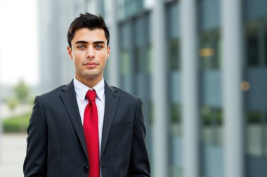 Sivri takım elbiseli ve kırmızı kravatlı kendine güvenen genç işadamı modern ofis binasının önünde poz veriyor, profesyonellik ve şehir planına karşı hırs yayıyor