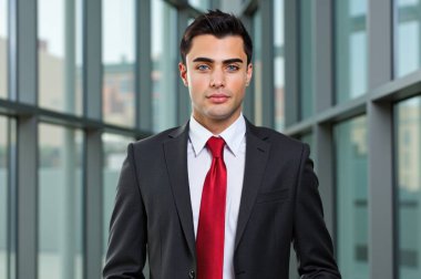 Keskin takım elbiseli genç işadamı modern ofis lobisinde duruyor, kararlılıkla pencereden dışarı bakıyor, arka planda şehir manzarası var. Azimli, başarılı kurumsal bireyin portresi