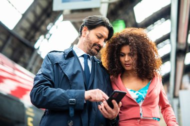 Takım elbiseli bir işadamı, tren istasyonunda iş konularını konuşurken akıllı telefonuyla bayan meslektaşına trenlerini beklerken bir şeyler gösteriyor.