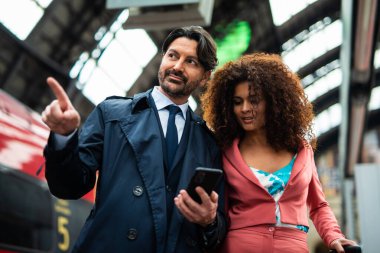 İş adamı bir eliyle bir tren istasyonunda dururken iş arkadaşına telefonunda bir şey gösteriyor.