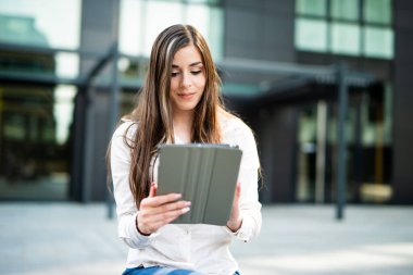 Dijital tablet kullanan genç bir iş kadını bankta oturuyor.