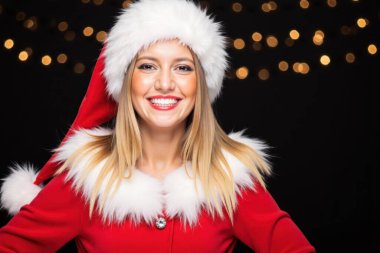 Noel Baba kostümlü neşeli sarışın kadın bulanık ışıklarla siyah bir arka plana karşı mutlu bir şekilde poz veriyor.