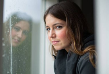 Genç bir kadın yağmurlu bir günde pencerenin kenarında duruyor, düşüncelere dalmış ve hayatı yansıtıyor, melankolik bir yalnızlık atmosferiyle çevrili.