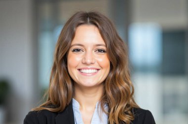 Uzun kahverengi saçlı genç iş kadını ofis ortamında gülümsüyor. O, siyah bir ceket ve mavi bir gömlek giyiyor.