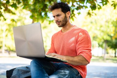 Güneşli bir park ortamında dizüstü bilgisayarda yazan odaklanmış serbest çalışan bir erkek..