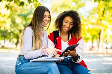 İki genç kadın, sonbahar yapraklarıyla çevrili açık havada çalışırken bir kitap ve tablet paylaşıyor.