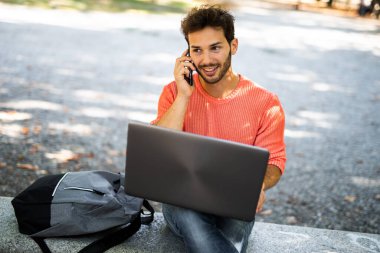 Neşeli, genç, erkek, güneşli bir parkta dizüstü bilgisayarı ve akıllı telefonu olan.