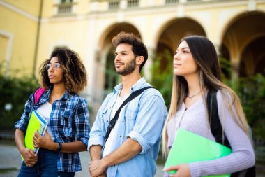 Farklı etnik kökenlere sahip üç genç öğrenci üniversite binalarının önünde yürüyüp sohbet ediyorlar.