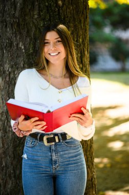 Genç öğrenci güneşli bir sonbahar gününde, huzurlu bir parkta mutlu bir şekilde ders çalışıyor, kendini bir ağacın altına atmış, kampüs hayatına ara verip, dinleniyor ve huzurlu atmosferin tadını çıkarıyor.