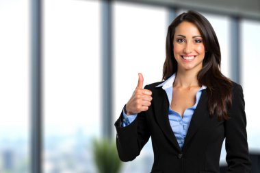 Profesyonel ofis ortamındaki mutlu iş kadını güven ve pozitiflik göstererek onay veriyor. Gülüşü başarıyı ve motivasyonu yansıtıyor. Arka planda bir şehir manzarası var.