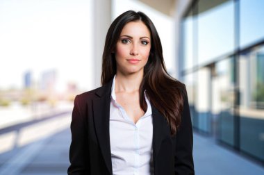 Genç bir iş kadını modern bir ofis binasının koridorunda poz veriyor. O siyah bir ceket ve beyaz bir gömlek giyiyor.