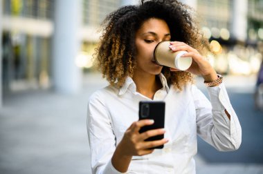 Güzel, genç, Afro-Amerikan bir yönetici kahve içerken akıllı telefonunu kullanıyor.