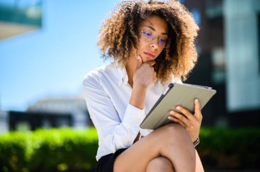 Genç iş kadını güneşli bir günde dışarıda oturuyor, tabletinden bir şeyler okuyor ve bu bilgiyi değerlendiriyor.