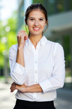 Ofis binasının önünde gülümseyen ve poz veren genç bir iş kadını, kollarını kavuşturmuş, beyaz gömlek ve siyah pantolon giyiyor.