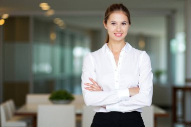 Modern bir ofiste kollarını kavuşturarak gülümseyen genç bir iş kadınının portresi, iş yerinde güven ve profesyonellik gösteriyor.