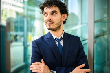 Takım elbiseli profesyonel bir genç, düşünceli bir kenara bakıyor, kollarını kavuşturmuş, şehir ortamında.