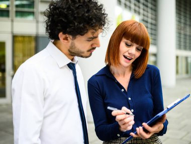 İki iş ortağı ofis dışında belgeleri tartışıyor, gülümsüyor ve modern gökdelenle işbirliği yapıyor arkalarında şehir manzarasını yansıtıyorlar.