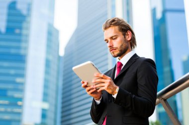 Keskin bir takım elbise içindeki odaklanmış genç işadamı hareketli bir şehrin silueti önünde bir tablet kullanıyor, modern bir şehir ortamında başarı ve verimlilik sergiliyor.