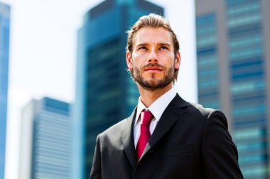 Kendine güvenen takım elbiseli işadamı modern şehir merkezinde kararlı ve hırslı görünüyor. Etrafı gökdelenler ve ofis binalarıyla çevrili. Başarının ve liderliğin portresi Umut ve ilhamla