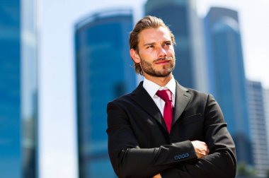 İş adamı, şehirdeki bazı gökdelenlerin önünde kollarını kavuşturmuş dururken gülümsüyor. Takım elbise ve kravat takıyor.