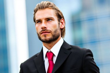 Kırmızı kravatlı genç işadamı, modern bir ofis binasının önünde duruyor, kararlı bir şekilde uzaklığa bakıyor, bir şehrin arka planında başarı ve hırsı somutlaştırıyor