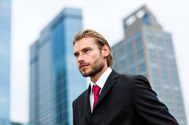 Sivri takım elbiseli genç işadamı gökdelenlerin arasında duruyor, modern şehir ortamında azim ve hırsla geleceğe bakıyor.