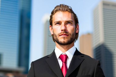 Keskin takım elbiseli kendine güvenen genç işadamı şehir gökyüzüne bakıyor, hırsı ve finans başarısını somutlaştırıyor. Şehir arkaplanı profesyonel atmosfere katkı sağlıyor. Parlak gün enerjiyi artırır.