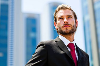 Keskin takım elbiseli kendine güvenen işadamı, yüksek binalarla çevrili şehir gökyüzüne iyimser bir şekilde bakıyor, başarıyı ve hırsı yansıtıyor.