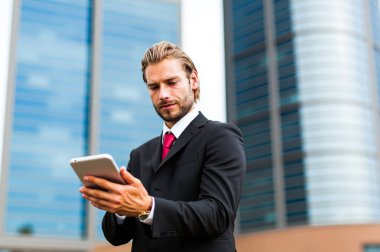 Sivri takım elbiseli kendine güvenen işadamı, şehir gökdelenleriyle çevrili, finans dünyasının modern yöneticiliğini somutlaştıran kurumsal binaların önünde tablete göz atıyor.