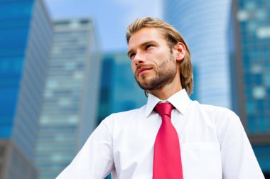 Modern ofis binalarının önünde duran beyaz gömlekli ve kırmızı kravatlı genç profesyonel ciddi bir ifadeyle başka yere bakıyor.