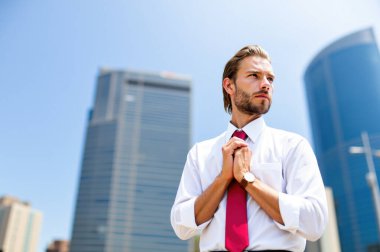 Genç profesyonel, kırmızı kravatını kendine güvenle düzeltiyor, hareketli bir şehir merkezinde modern gökdelenlerin önünde duruyor, hırs ve başarıyı somutlaştırıyor.