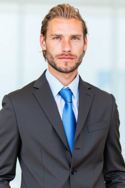 İş adamı kendine güveni ve profesyonelliği yayıyor, parlak bir ofis ortamında duruyor, şık bir takım elbise ve kravat takıyor.