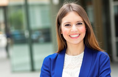 Modern bir şehir ortamında özgüvenle gülümseyen bir iş kadınının portresi profesyonelliği ve başarıyı gözler önüne seriyor.